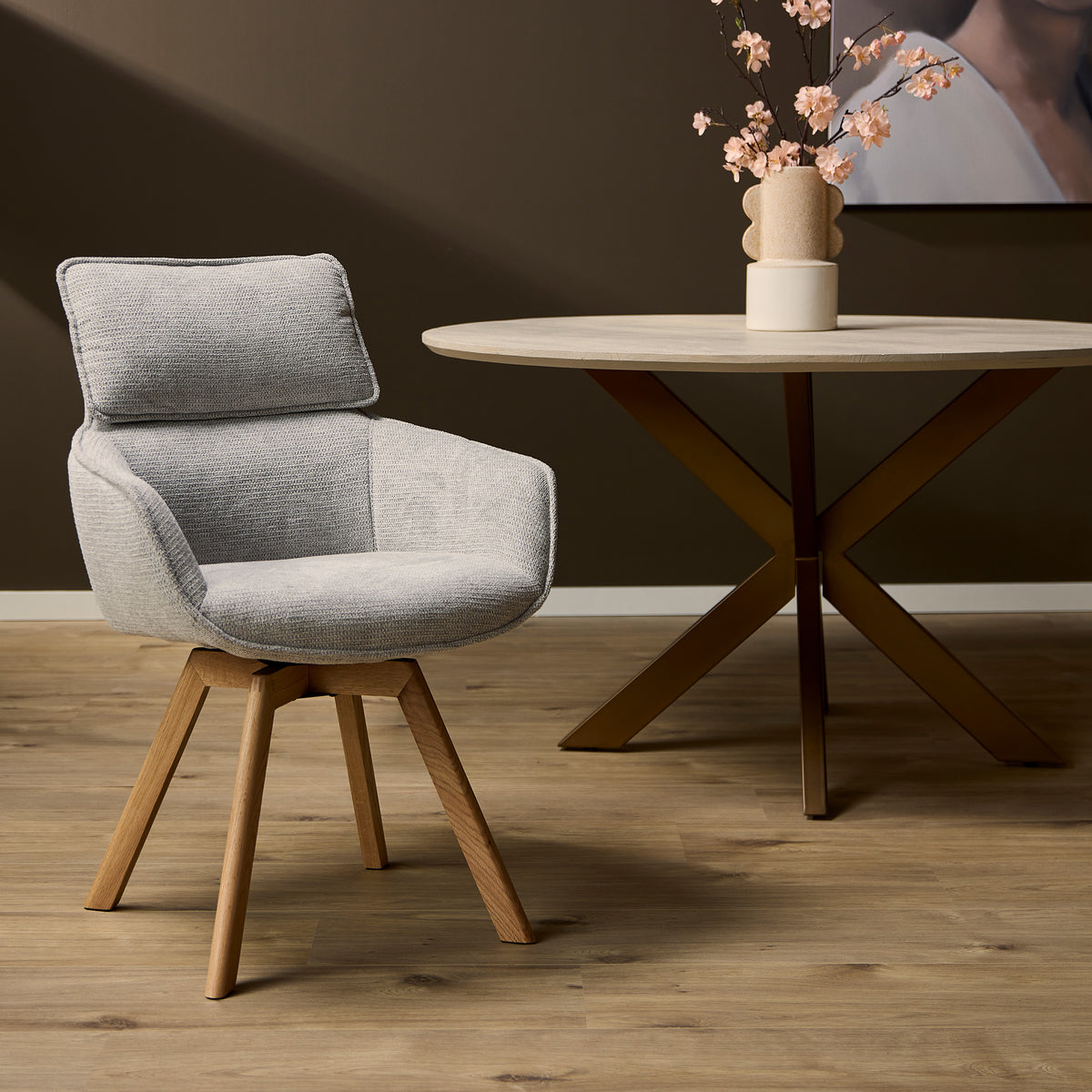 Eetkamerstoel Jona in taupe stof, ronde rug en armleuningen, houten pootjes.