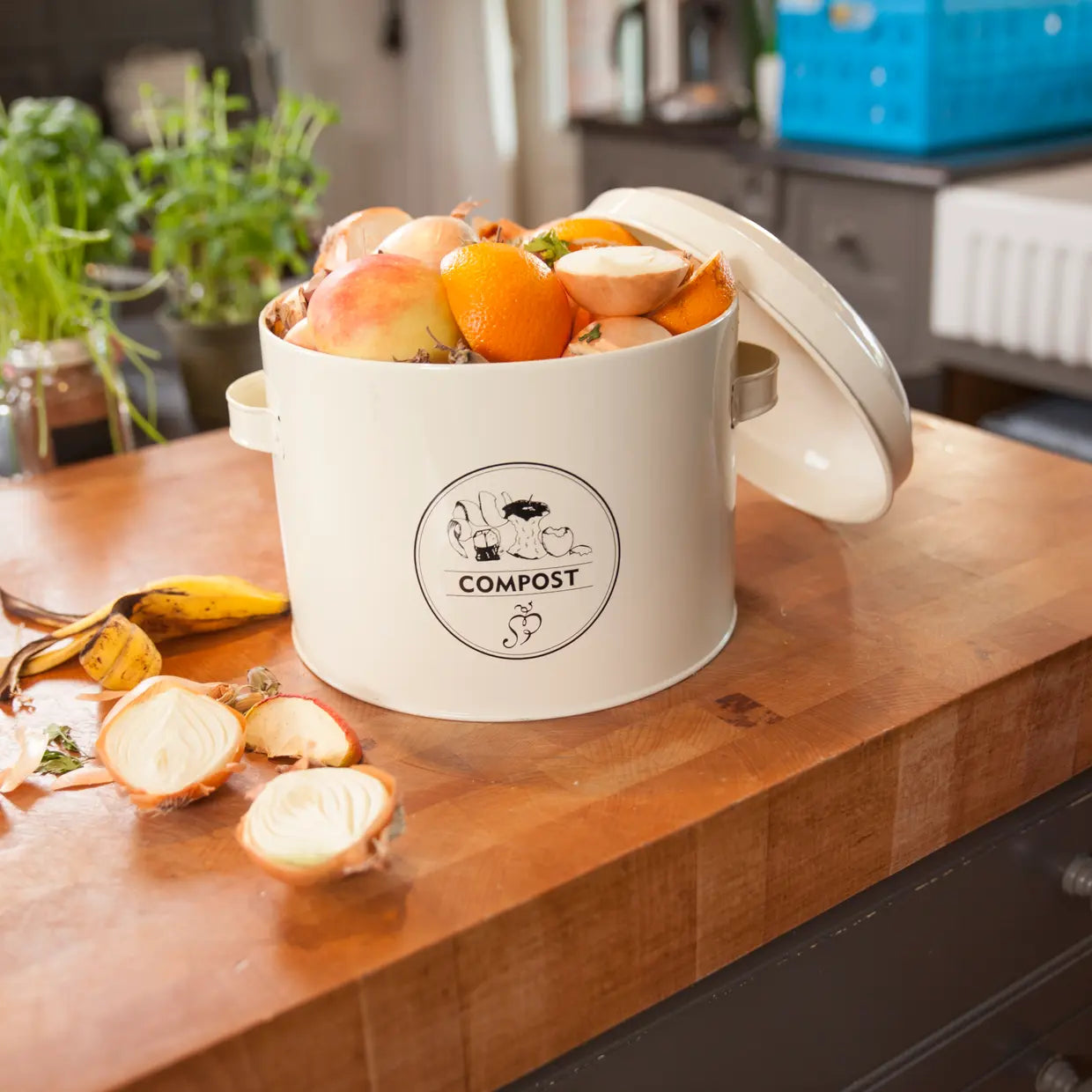 Keukencomposter in crème met open deksel en COMPOST-logo, gevuld met etensresten